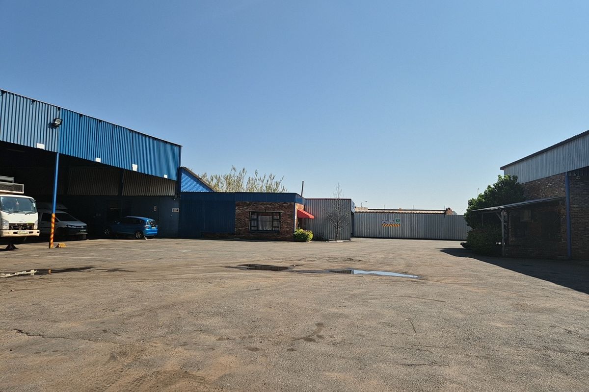 Spacious industrial yard featuring corrugated metal warehouse structures, brick facade sections, and ample open-air parking space.
