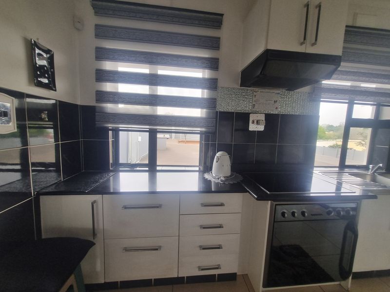 A functional kitchen featuring white cabinetry, black granite countertops, a built-in oven and hob, and a tiled backsplash with modern roller blinds.