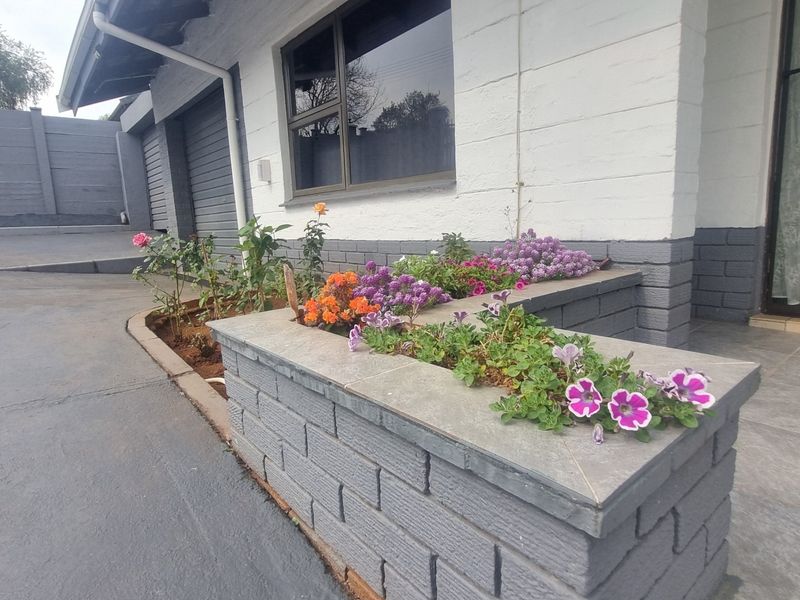 Front exterior view featuring a paved driveway, garage access, and a well-maintained brick planter box with established garden beds.
