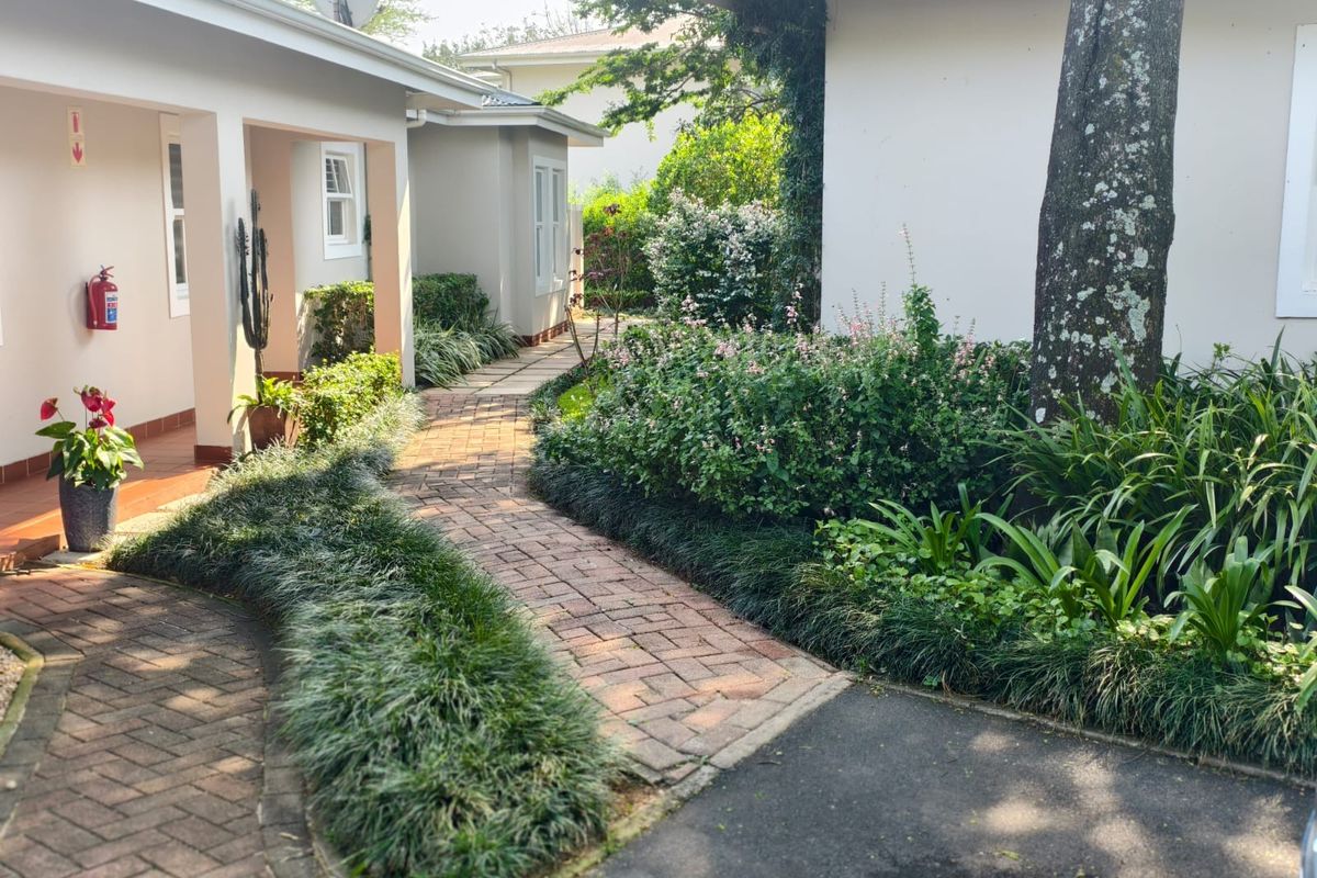 A well-maintained exterior view featuring a brick paved pathway leading through a landscaped garden between townhouse units with a covered patio area.