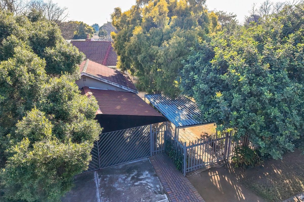 Aerial view of the property showing the roof structure, carport, metal security fencing, and paved entrance walkway surrounded by mature trees.