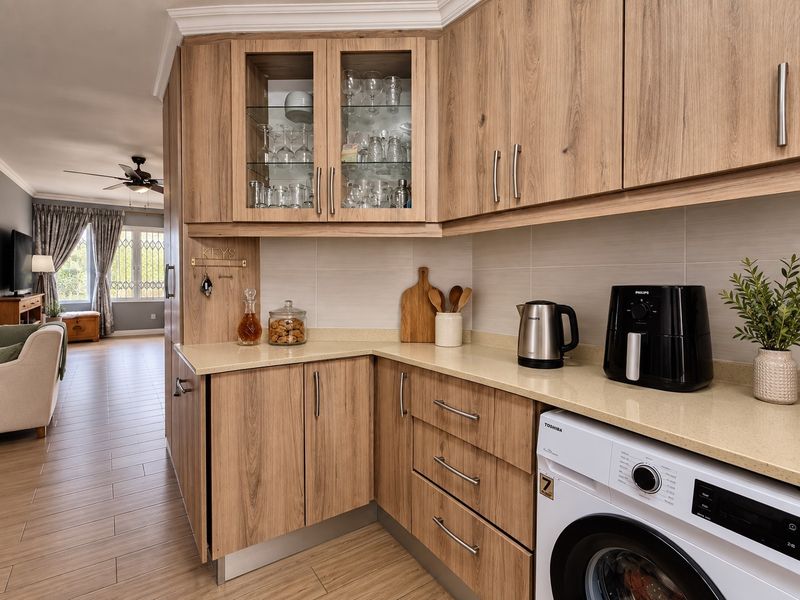 An open-plan kitchen area featuring modern wood-look cabinetry, stone countertops, and an integrated washing machine, flowing into a bright living space.