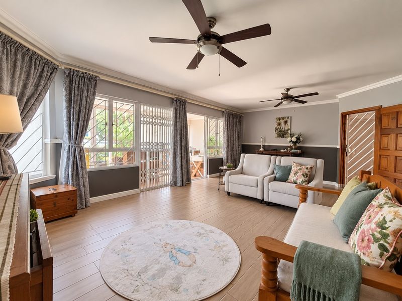 Spacious living room featuring tiled flooring, dual ceiling fans, and large windows with security gates, providing access to an outdoor patio area.