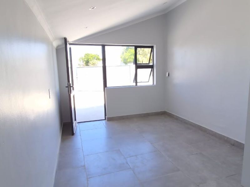 A bright, empty bedroom featuring neutral tiled flooring, recessed lighting, and direct access to an outdoor area through a glass-paneled door.