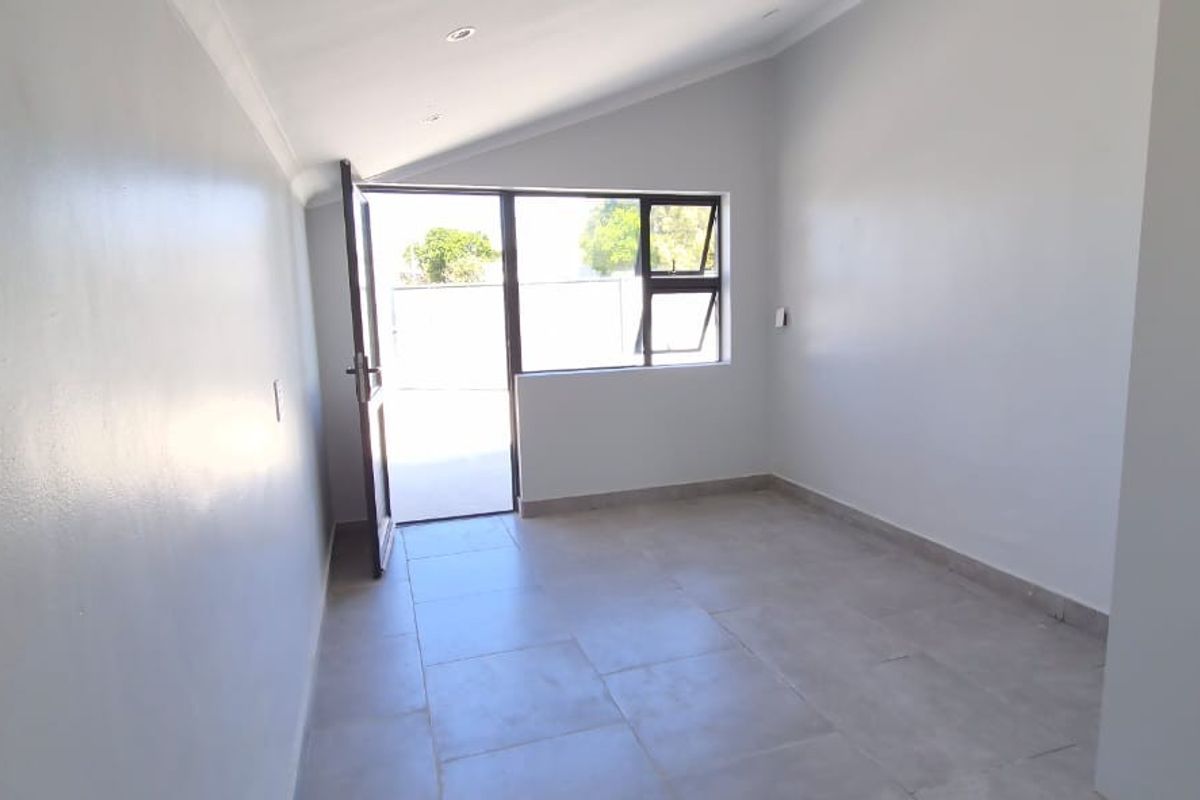 A bright, empty bedroom featuring neutral tiled flooring, recessed lighting, and direct access to an outdoor area through a glass-paneled door.