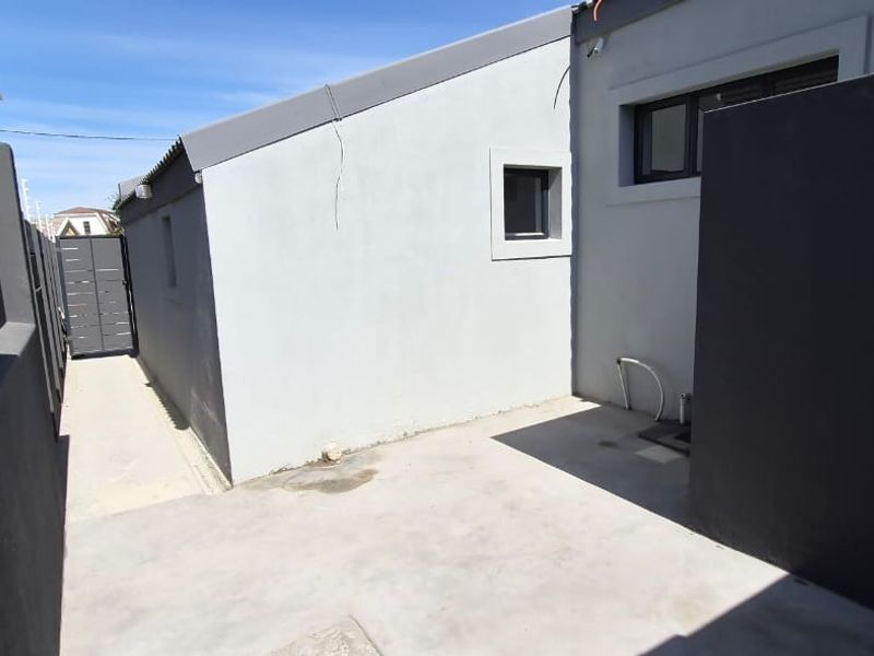 A paved rear exterior area featuring a concrete walkway, boundary walls, and external plumbing fixtures along the side of the building.