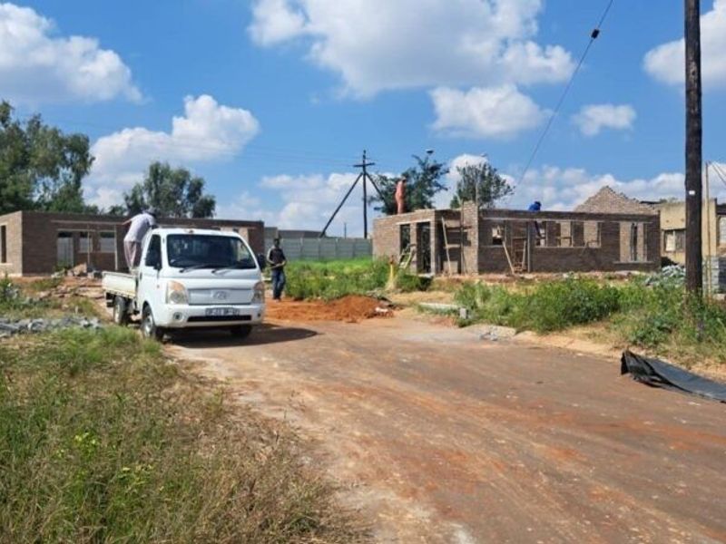 A construction site showing partially built brick structures and an unpaved access road in a developing residential area.