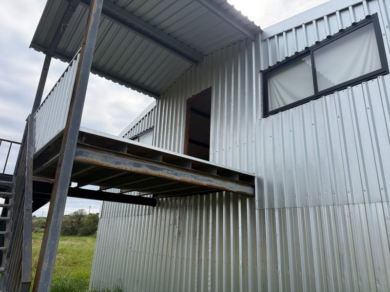 Exterior view of a corrugated metal structure featuring a raised steel balcony, external staircase, and black aluminium window frames.