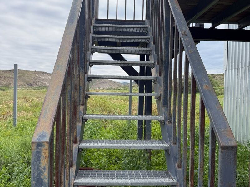 An exterior view of an elevated metal staircase leading to a structure, showing signs of surface rust and weathering.