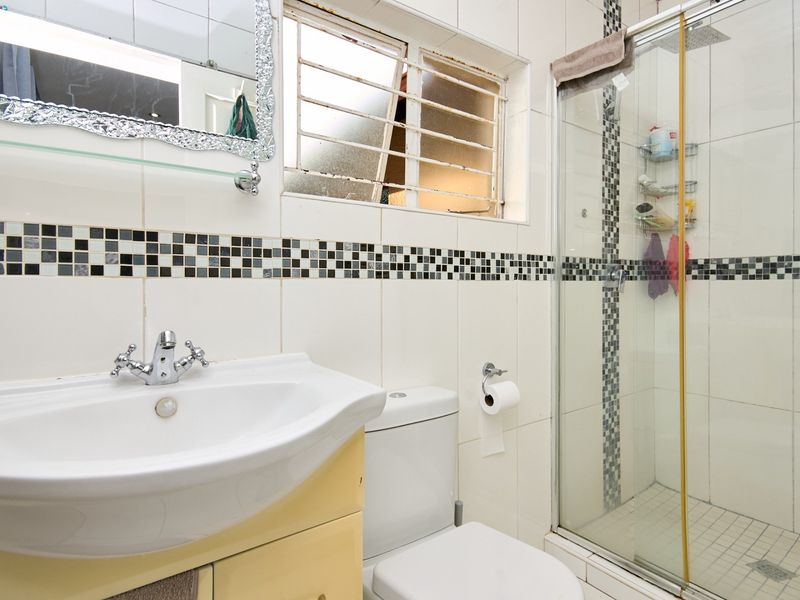 A functional bathroom featuring a pedestal vanity, toilet, and a glass-enclosed shower with decorative mosaic tile accents and security-barred window.