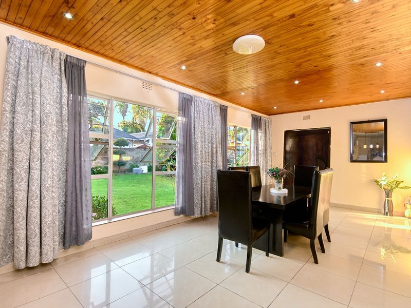 A bright dining room featuring polished tiled floors, a warm timber-paneled ceiling with recessed lighting, and large windows overlooking the garden.