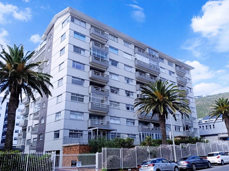 A multi-story apartment building featuring a white facade, multiple balconies, and secure perimeter fencing with palm trees in the foreground.