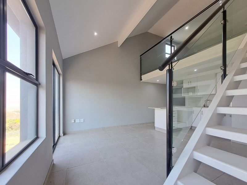 Modern open-plan living area featuring a staircase with glass balustrade, vaulted ceilings, tiled floors, and a sleek kitchen with built-in cabinetry.
