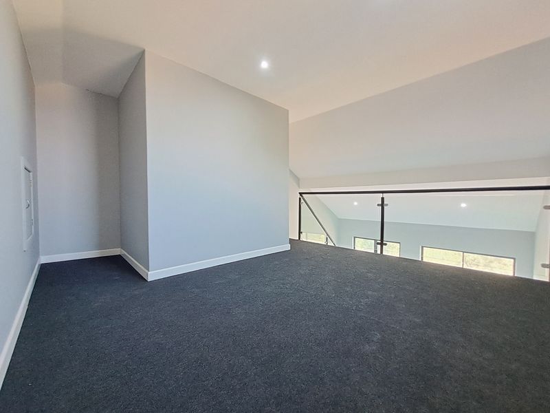 A spacious, empty mezzanine bedroom area featuring dark carpeting, a modern glass balustrade, and recessed downlighting, overlooking the lower level.