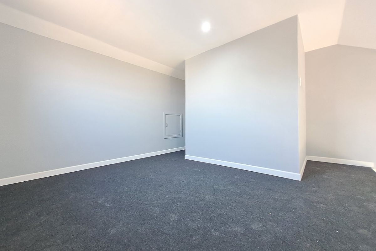 A clean, empty bedroom featuring neutral grey walls, dark carpeting, and a small wall-mounted access hatch.