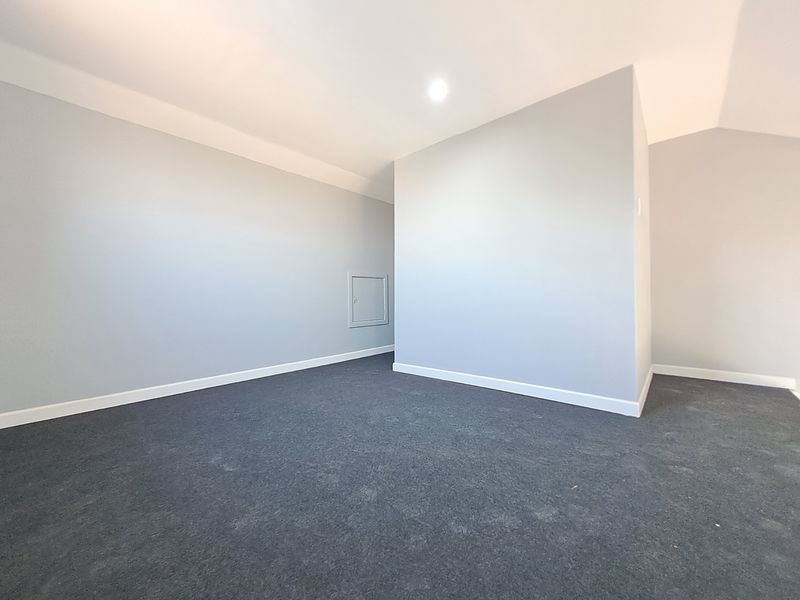 A clean, empty bedroom featuring neutral grey walls, dark carpeting, and a small wall-mounted access hatch.
