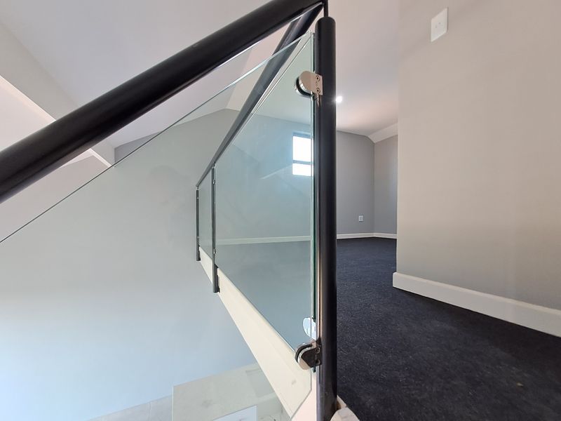 A modern hallway landing featuring a sleek glass balustrade with a black metal handrail and dark carpeted flooring.