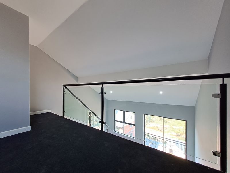 A bright mezzanine hallway featuring a modern glass balustrade, vaulted ceilings, and recessed lighting, overlooking the living area below.