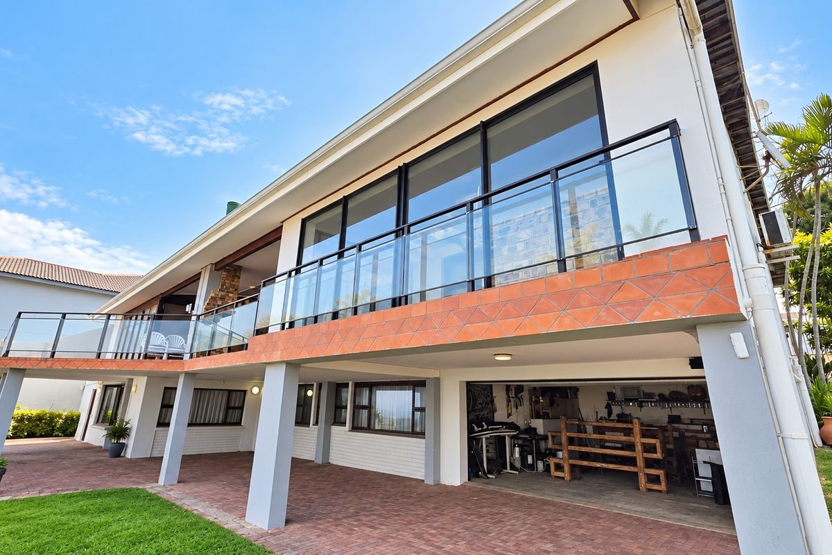 Rear elevation featuring a spacious paved patio, a glass-balustraded balcony, and a ground-level garage area with integrated structural columns.