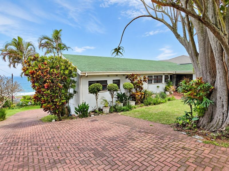 Front exterior view of a single-story house featuring a brick paved driveway, green pitched roof, and established garden with ocean views.