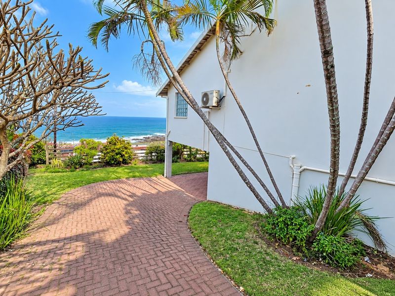 A well-maintained rear exterior view featuring a paved walkway, landscaped garden, and ocean views, with an external air conditioning unit mounted on the wall.