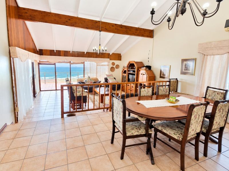 Spacious open-plan living and dining area featuring vaulted ceilings with exposed timber beams, ceramic tiling, and direct access to a balcony with ocean views.