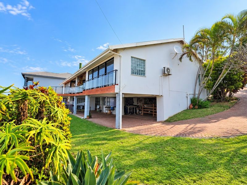 Rear view of the property featuring a covered patio area, glass balustrade balcony, paved driveway, and established garden surroundings.