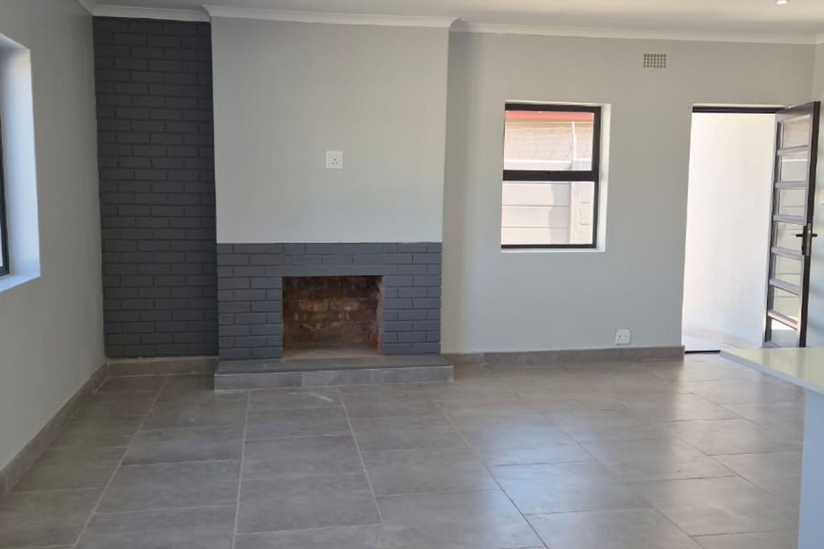 A bright, open-plan living area featuring neutral grey walls, tiled flooring, and a prominent brick fireplace, offering a clean and modern blank canvas.