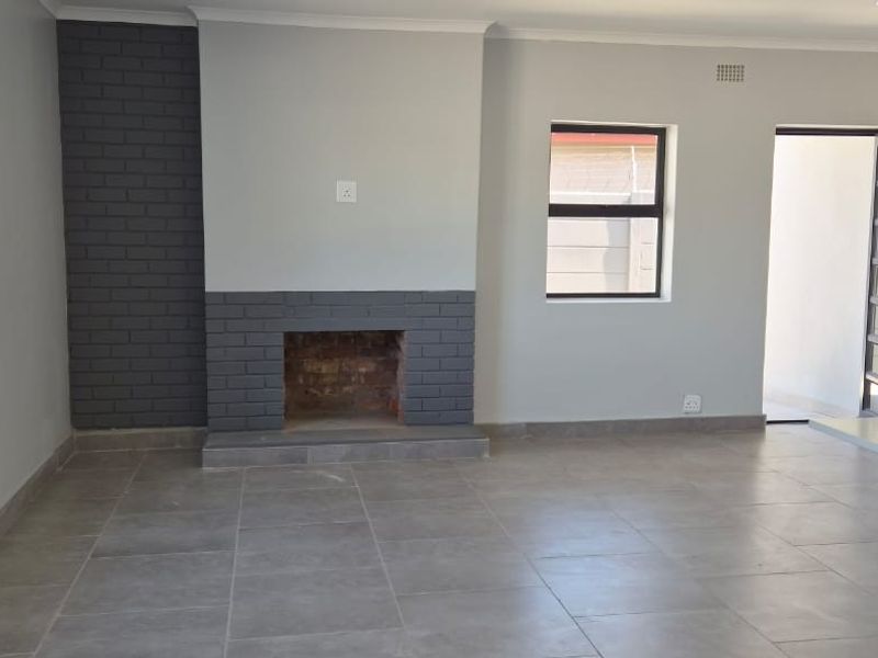A bright, open-plan living area featuring neutral grey walls, tiled flooring, and a prominent brick fireplace, offering a clean and modern blank canvas.