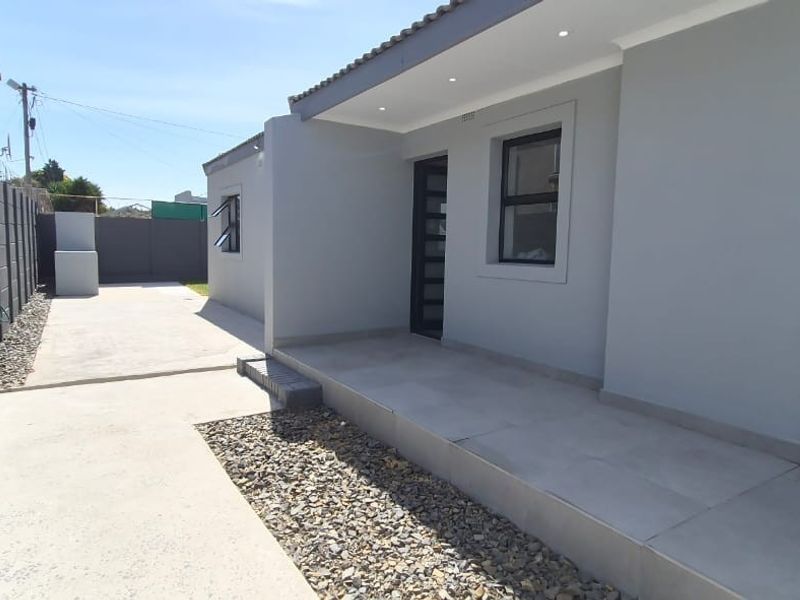 A well-maintained rear exterior view featuring a tiled patio, concrete driveway, and modern grey facade with recessed lighting and secure boundary walling.