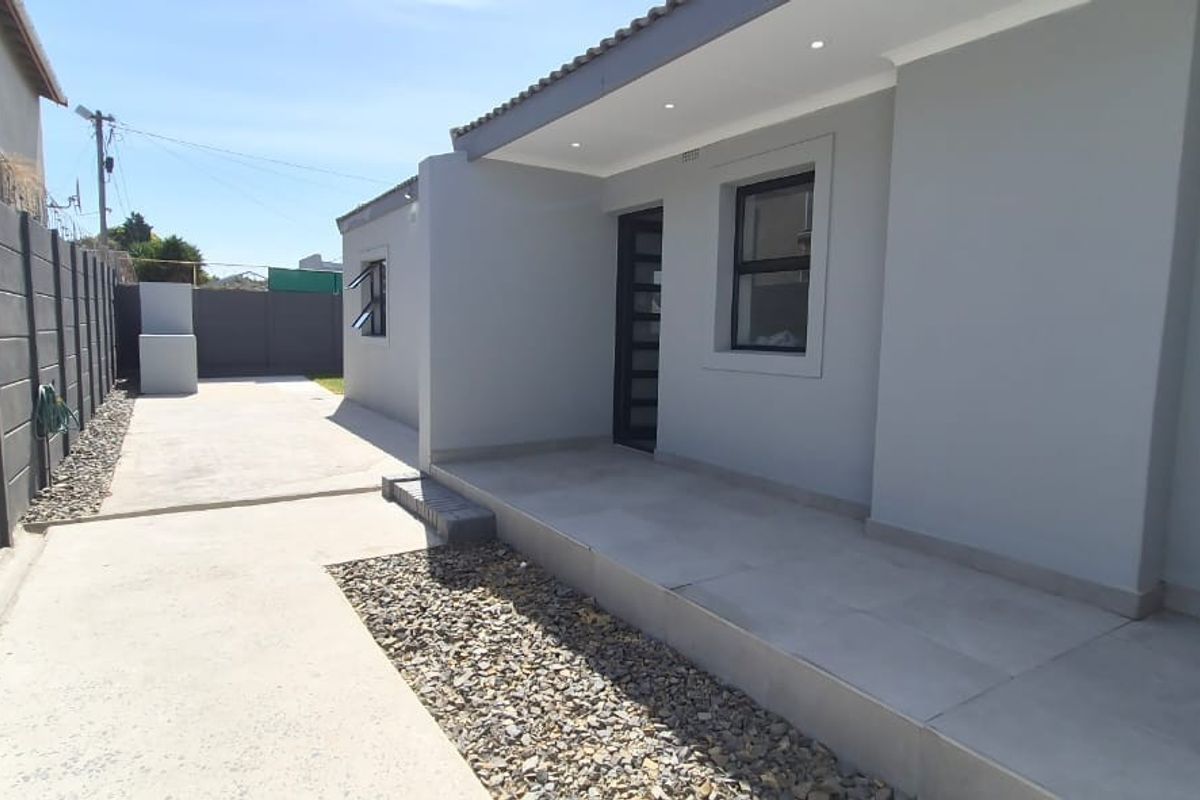 A well-maintained rear exterior view featuring a tiled patio, concrete driveway, and modern grey facade with recessed lighting and secure boundary walling.