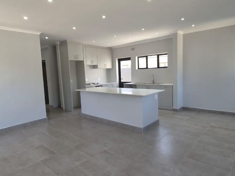 A spacious, modern open-plan kitchen and living area featuring neutral tones, tiled flooring, a central island, and contemporary built-in cabinetry.