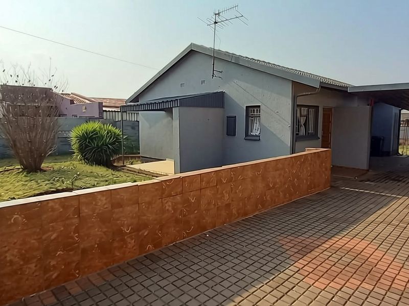 Front exterior view of a single-story house featuring a paved driveway, attached carport, and a tiled boundary wall with a small garden area.