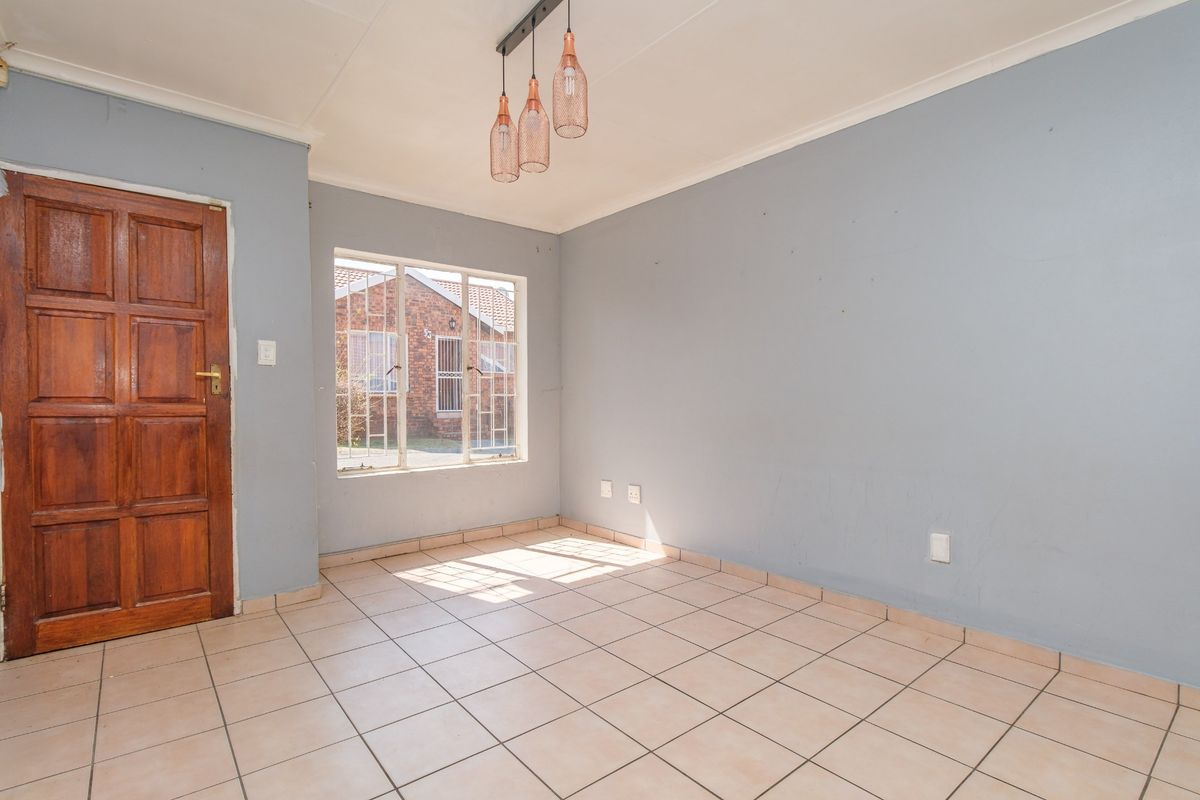 A bright, empty living area featuring neutral tiled flooring, a solid timber door, and natural light through a window with security bars.