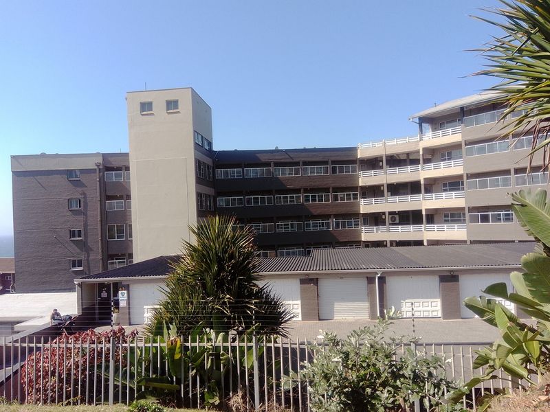 Exterior view of a multi-story apartment complex featuring a brick facade, multiple balconies, and ground-level garage units with secure perimeter fencing.