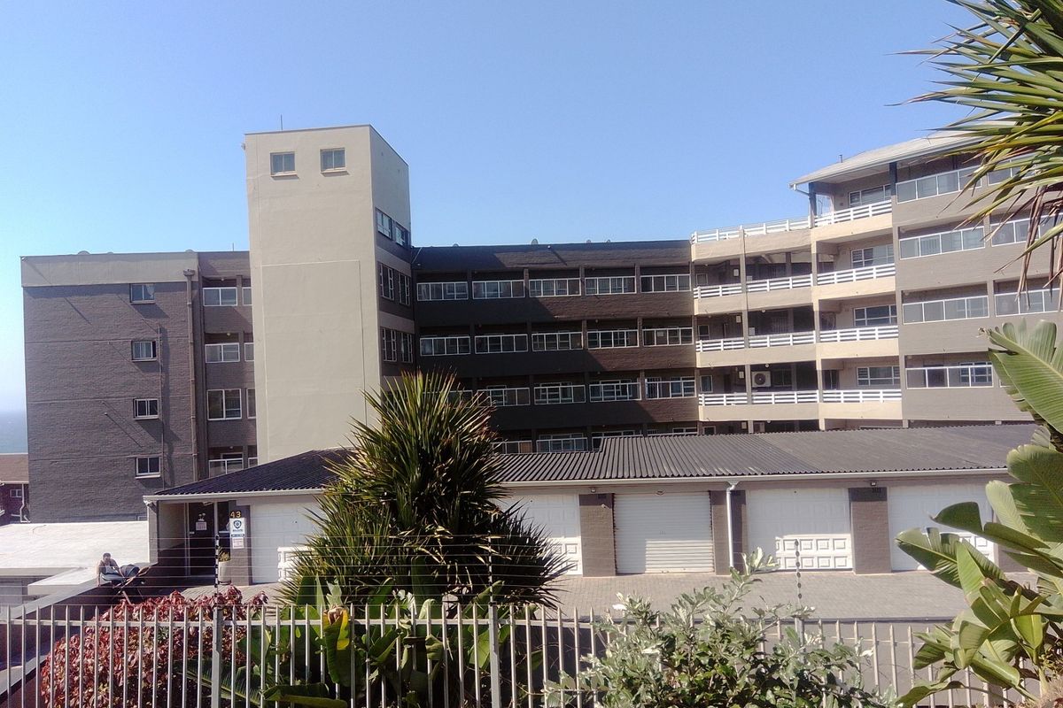 Exterior view of a multi-story apartment complex featuring a brick facade, multiple balconies, and ground-level garage units with secure perimeter fencing.