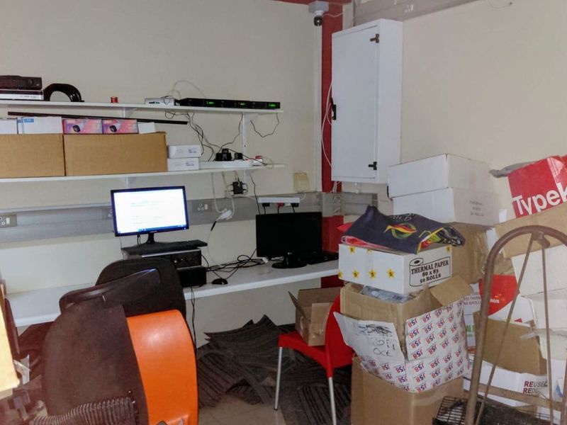 A utility or server room featuring built-in desk space, wall-mounted shelving, and an electrical distribution board, currently used for storage and equipment housing.