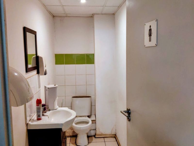 A functional commercial restroom featuring a toilet, washbasin, wall-mounted hand dryer, and tiled splashback, requiring cosmetic updates to improve its overall condition.