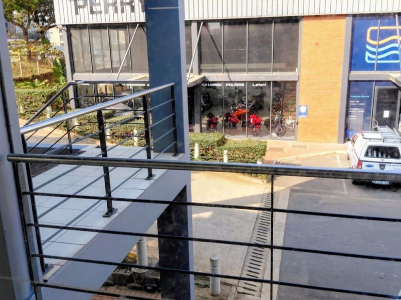 Elevated view of a commercial property exterior featuring a tiled balcony with metal railings overlooking a paved parking area and glass-fronted retail units.