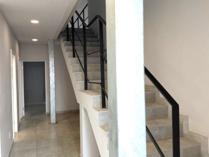 A clean, modern hallway featuring a tiled staircase with a black metal handrail, recessed lighting, and neutral finishes throughout the industrial space.