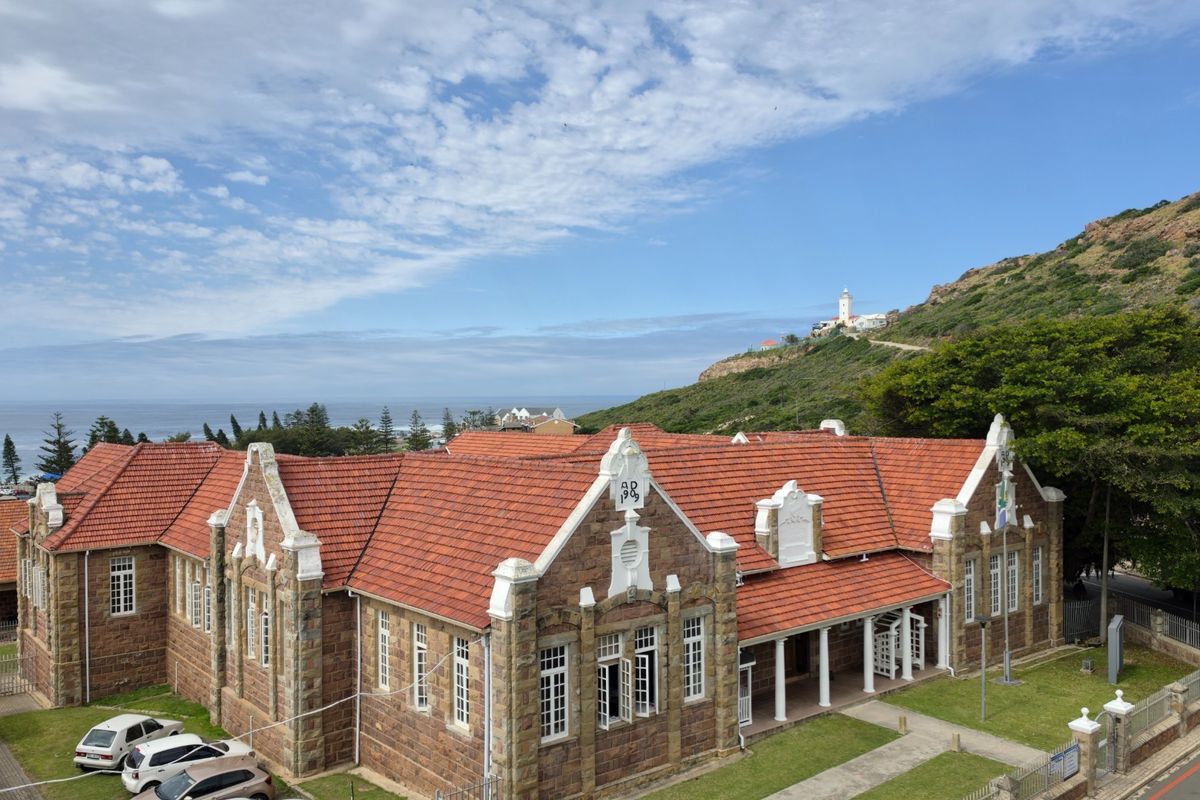 Aerial view of a historic stone building featuring a terracotta tile roof, gabled architecture, and a covered veranda, set against a coastal landscape.