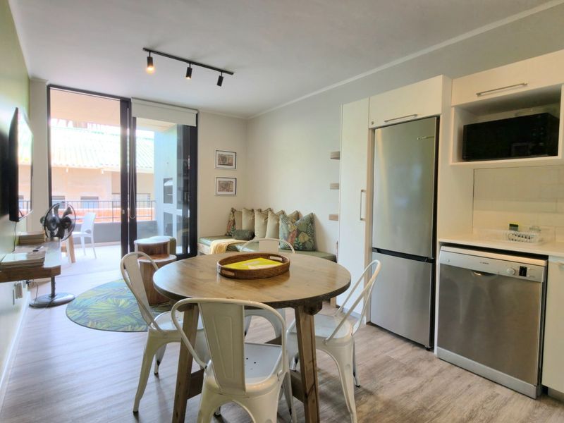 Modern open-plan apartment featuring a compact kitchen with integrated appliances, dining area, and direct access to a private balcony through sliding glass doors.