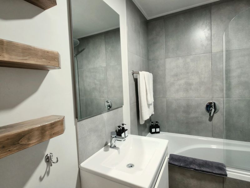 Modern bathroom featuring a vanity basin, wall-mounted mirror, built-in bathtub with a glass shower screen, and contemporary grey wall tiling.