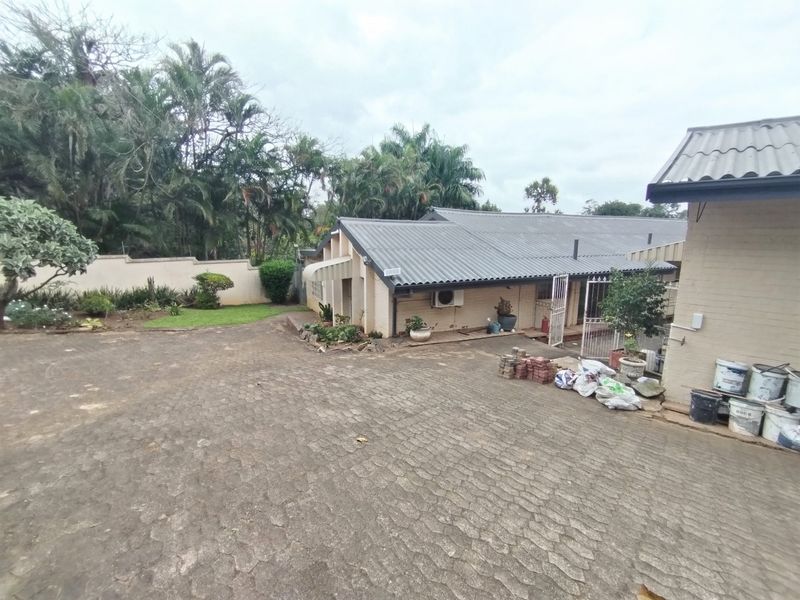 Rear exterior view of the property featuring a large brick-paved driveway, corrugated metal roofing, and a perimeter boundary wall.