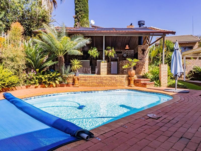 A well-maintained outdoor pool area featuring brick paving, a covered patio with a built-in fireplace, and established garden landscaping.