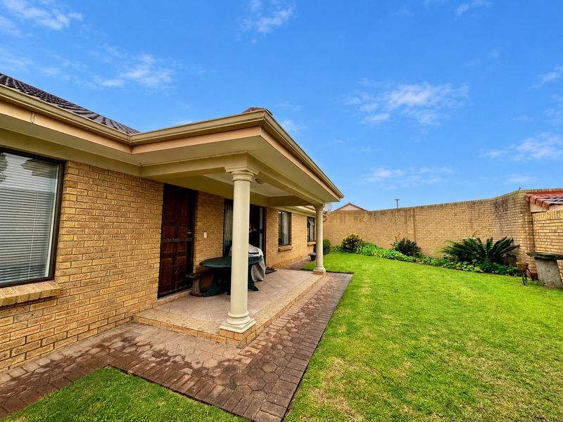 A well-maintained rear exterior featuring a covered patio with classical columns, brick facade, paved walkway, and a private lawn area enclosed by brick walls.