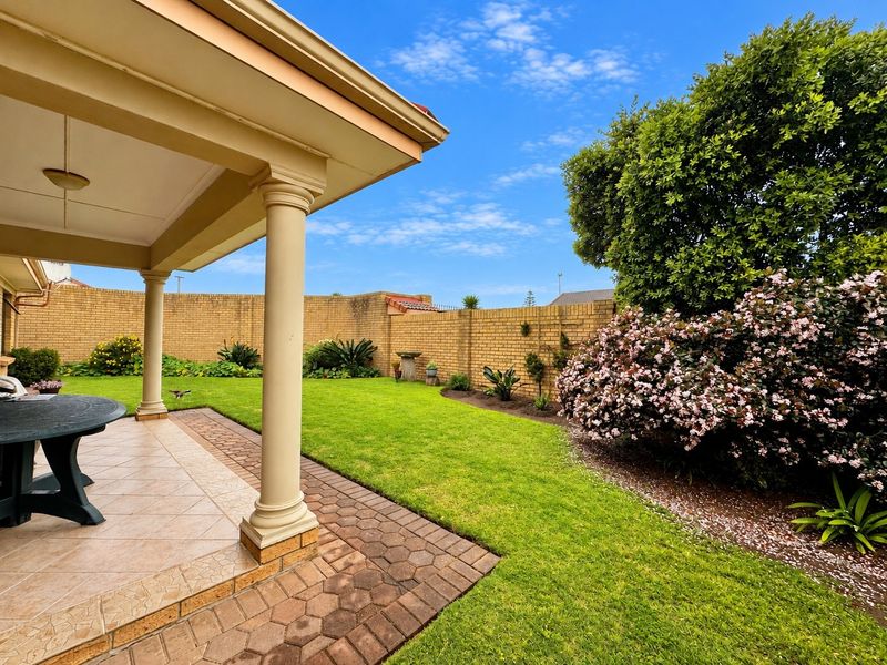 A well-maintained covered patio featuring classical columns, overlooking a lush green garden with a paved walkway and secure brick boundary wall.