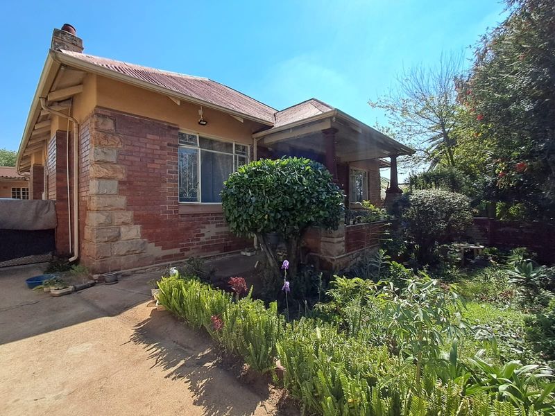 Front elevation of a traditional brick house featuring stone quoins, a covered porch, and a corrugated metal roof, set within a mature garden.