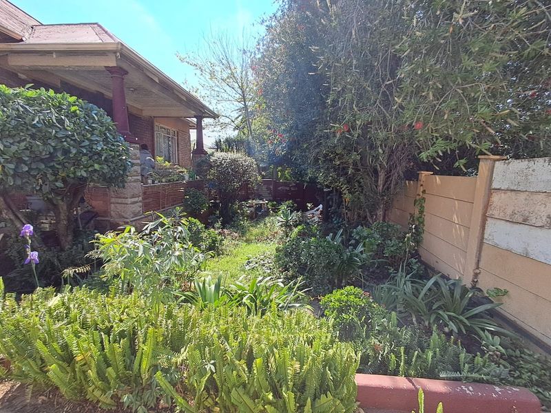 A lush, established garden featuring a variety of mature plants, a precast concrete boundary wall, and access to a covered veranda with brick pillars.