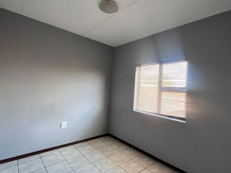 A clean, empty bedroom featuring neutral grey walls, tiled flooring, and a window fitted with venetian blinds.
